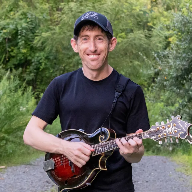 Greg Goodson smiling while holding his mandolin on a wooded trail